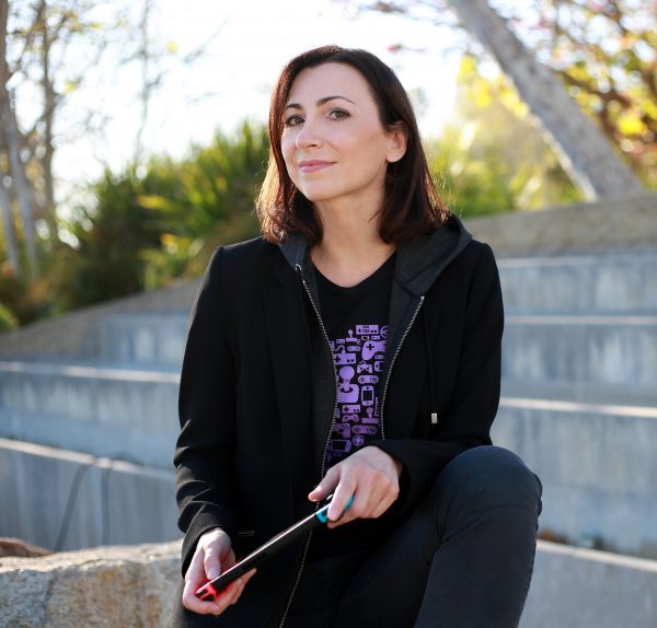 Photo of Celia Hodent, a white woman with brown hair, wearing a purple video game tshirt and a black jacket. She's sitting outside on concrete stairs and is looking at the camera, smiling.